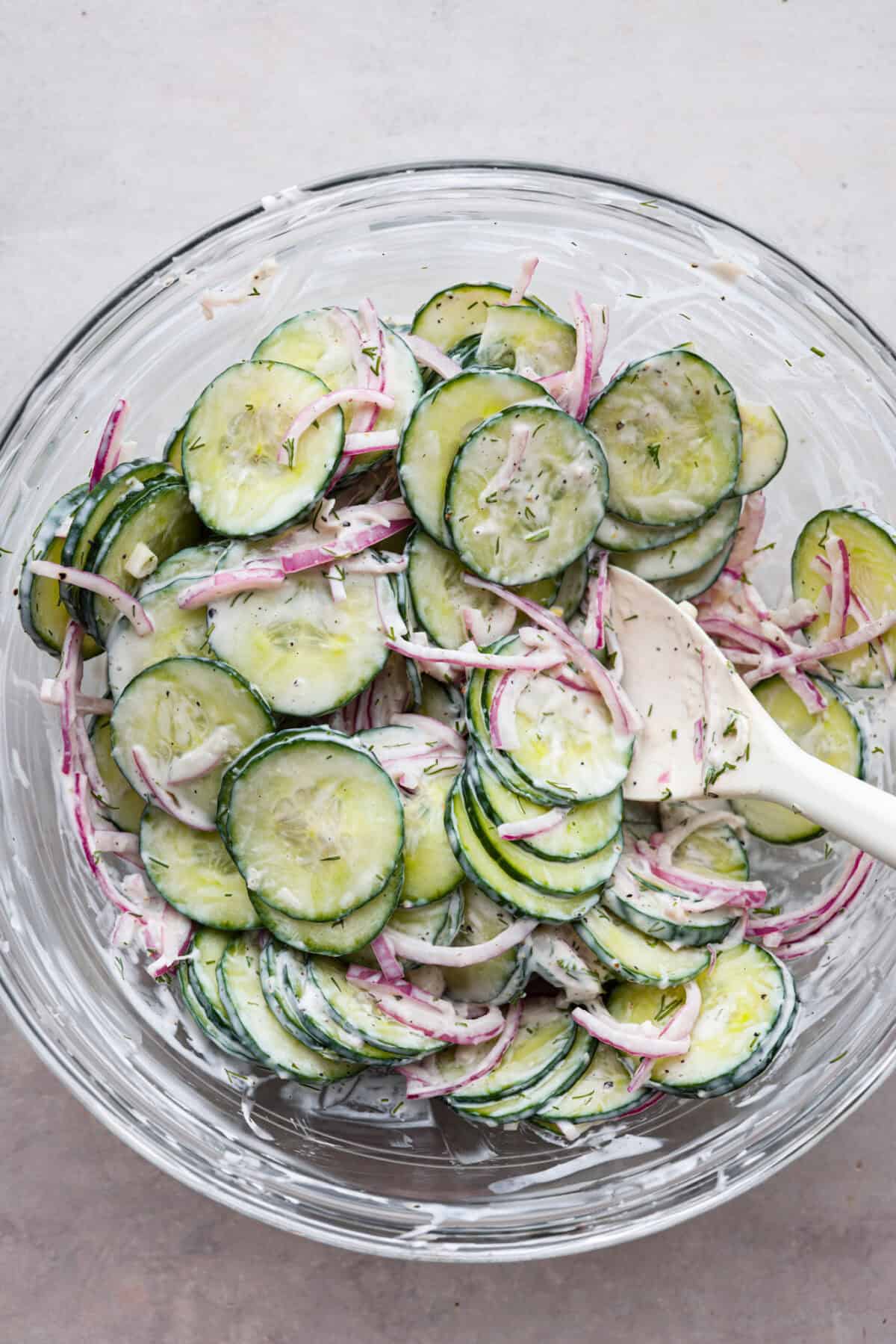 All of the ingredients for creamy cucumber salad being stirred in a glass bowl. 