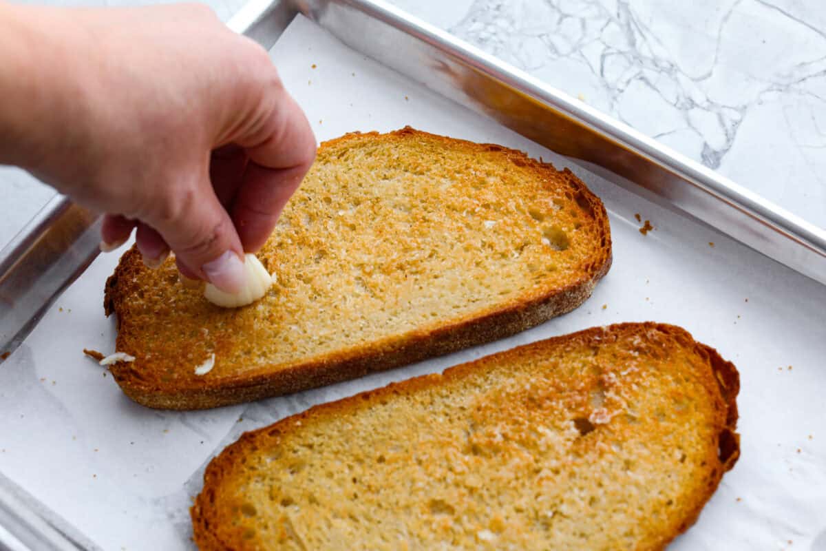 Overhead shot of someone rubbing garlic clove on top of toasted bread. 