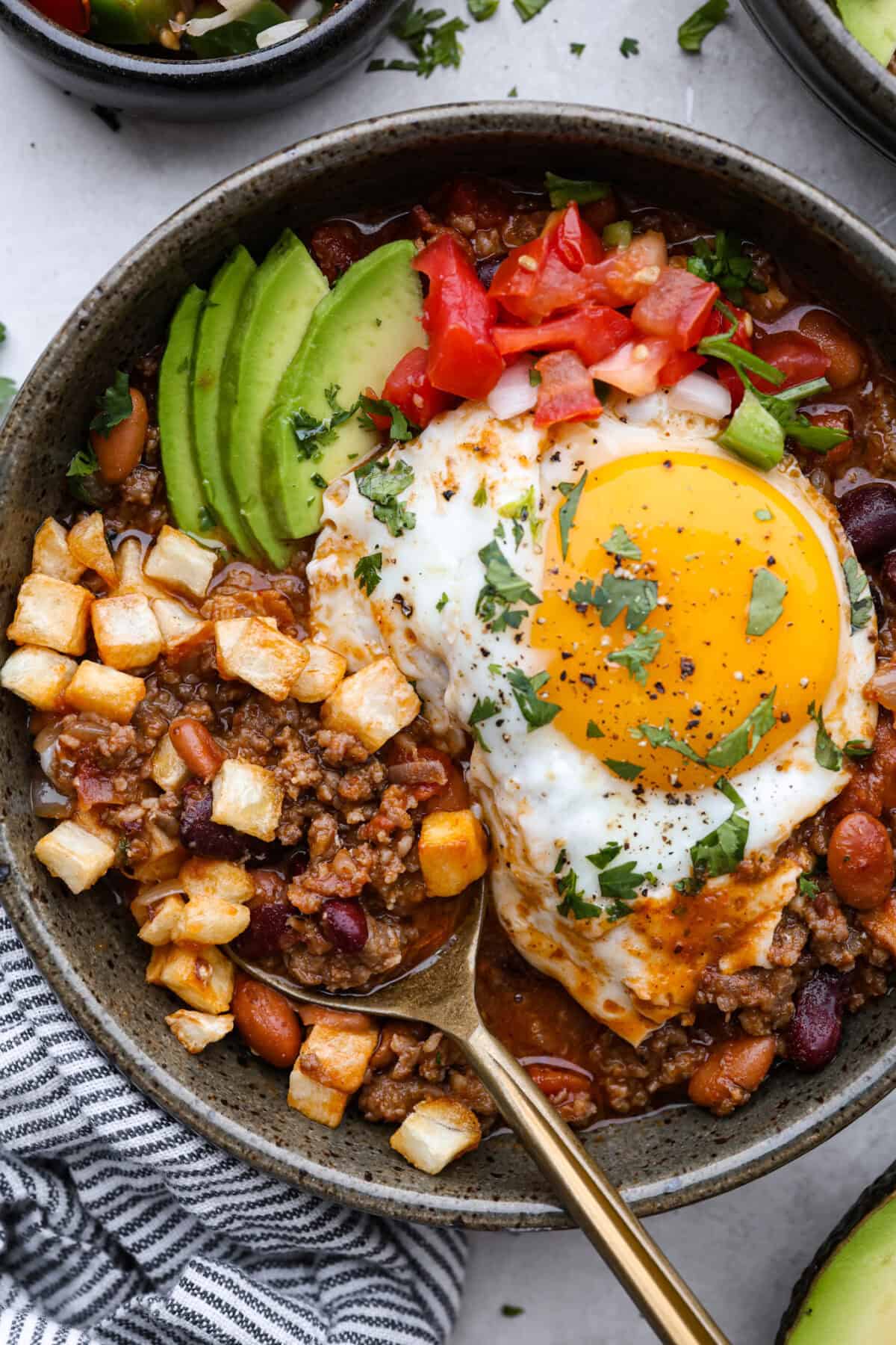 Overhead shot of plated breakfast chili with an egg, pico de gallo, cilantro, avocado and hash browns on top. 