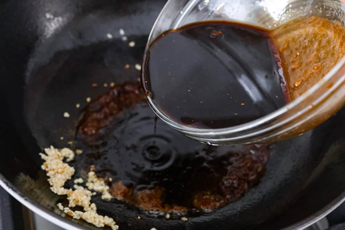 Overhead shot of someone pouring teriyaki sauce into the skillet with oil and garlic. 