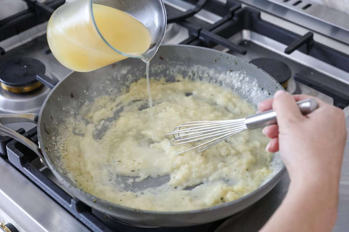 Angle shot of someone pouring the chicken broth, half and half, and lemon juice in the the butter and flour mixture. 