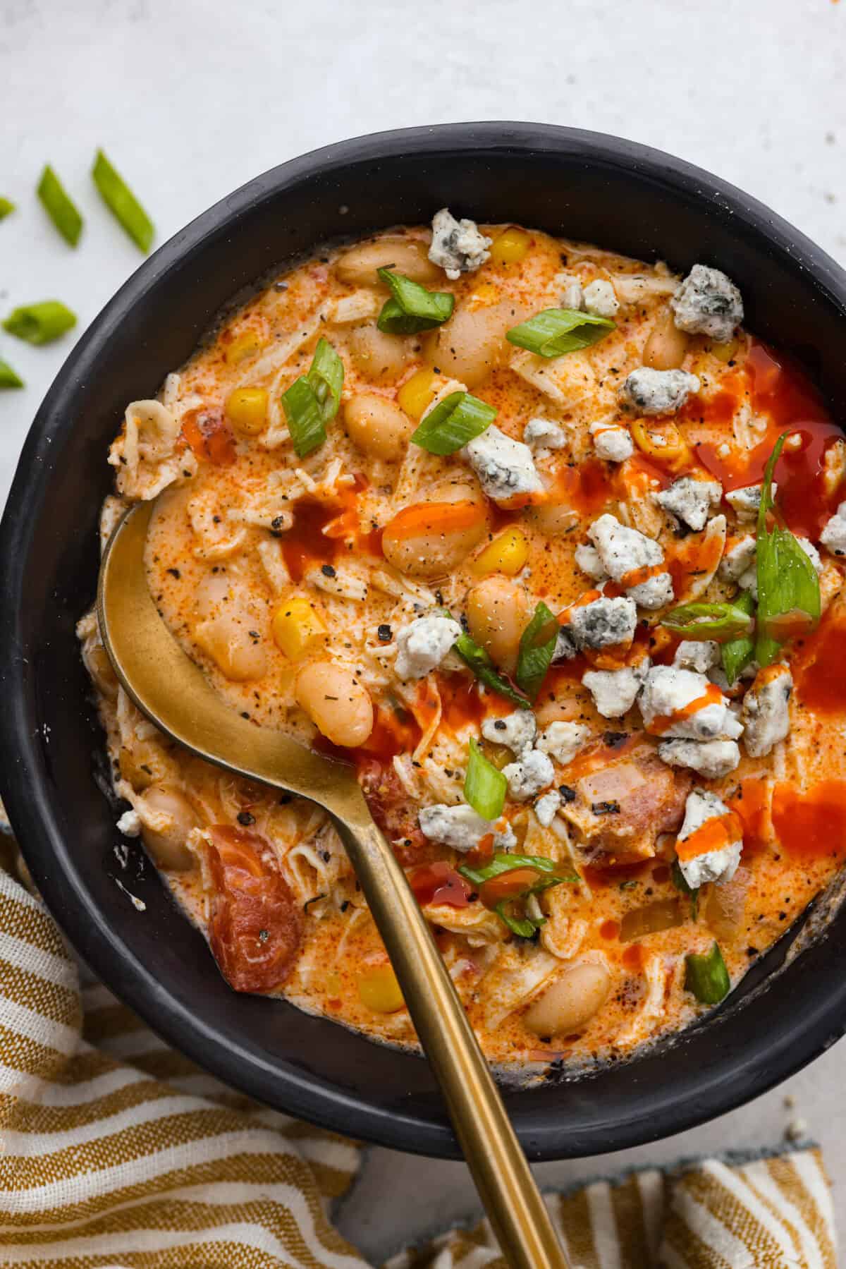 Overhead shot of a bowl of crockpot buffalo chicken chili with a gold spoon, topped with green onion, blue cheese crumbles and a dash more hot suace. 