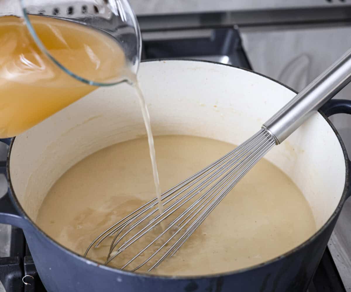 Angle shot of someone pouring in chicken broth into the roux.