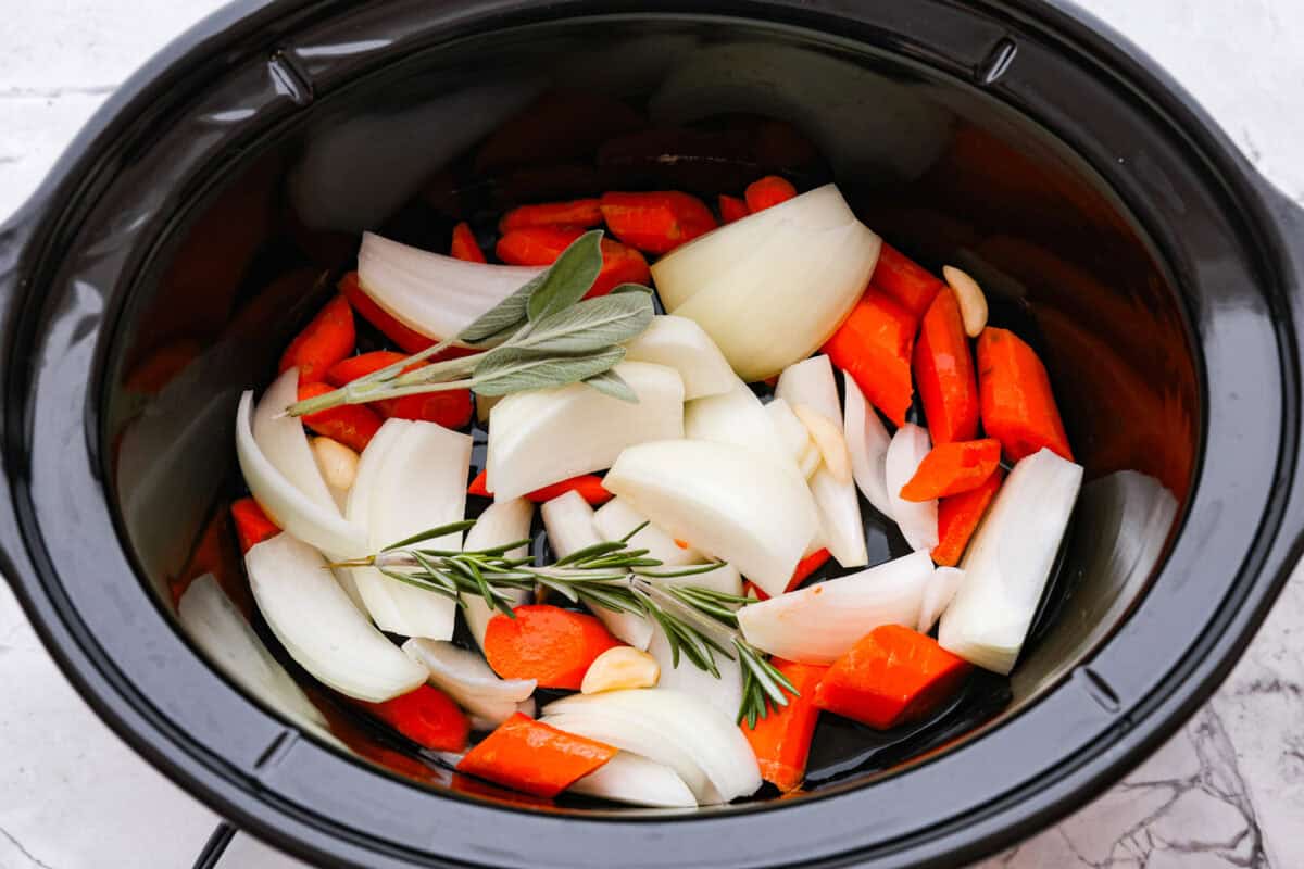 Overhead shot of onion, carrots, garlic and sprigs of herbs in the bottom of the crockpot. 