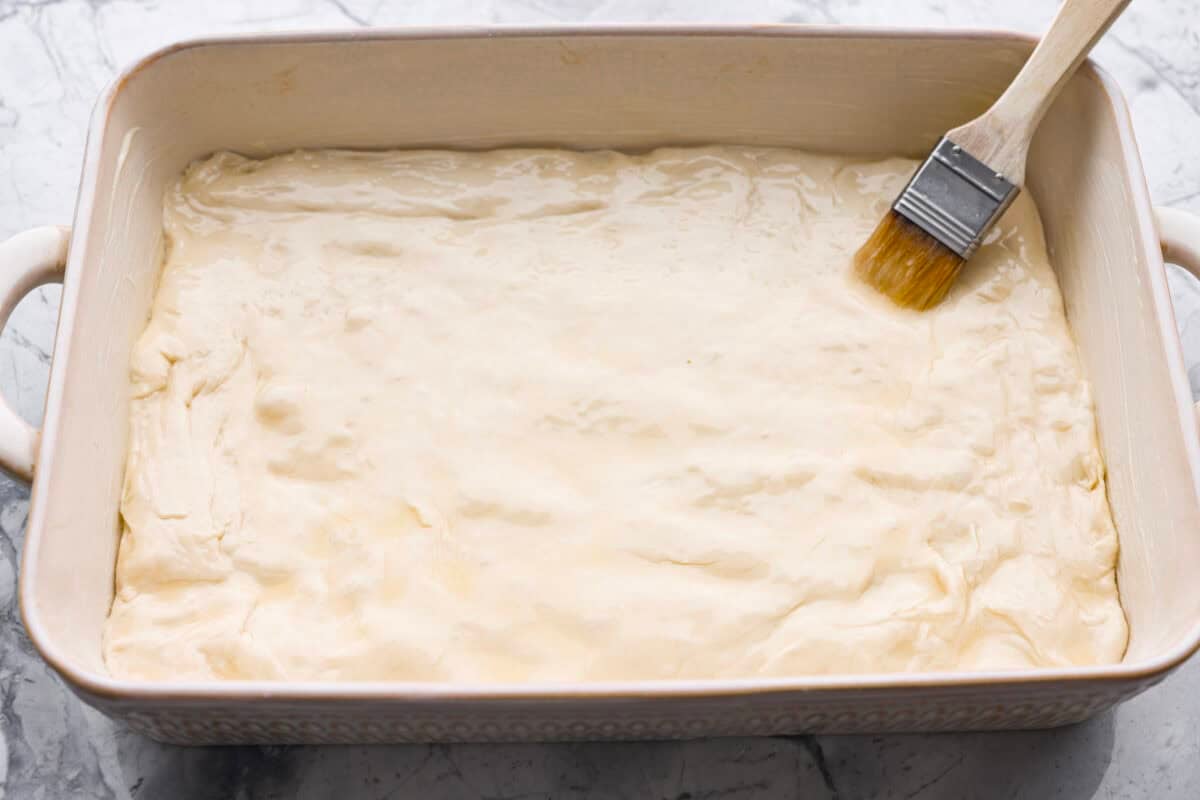 Someone brushing melted butter over the dough in a 9x13 baking dish. 