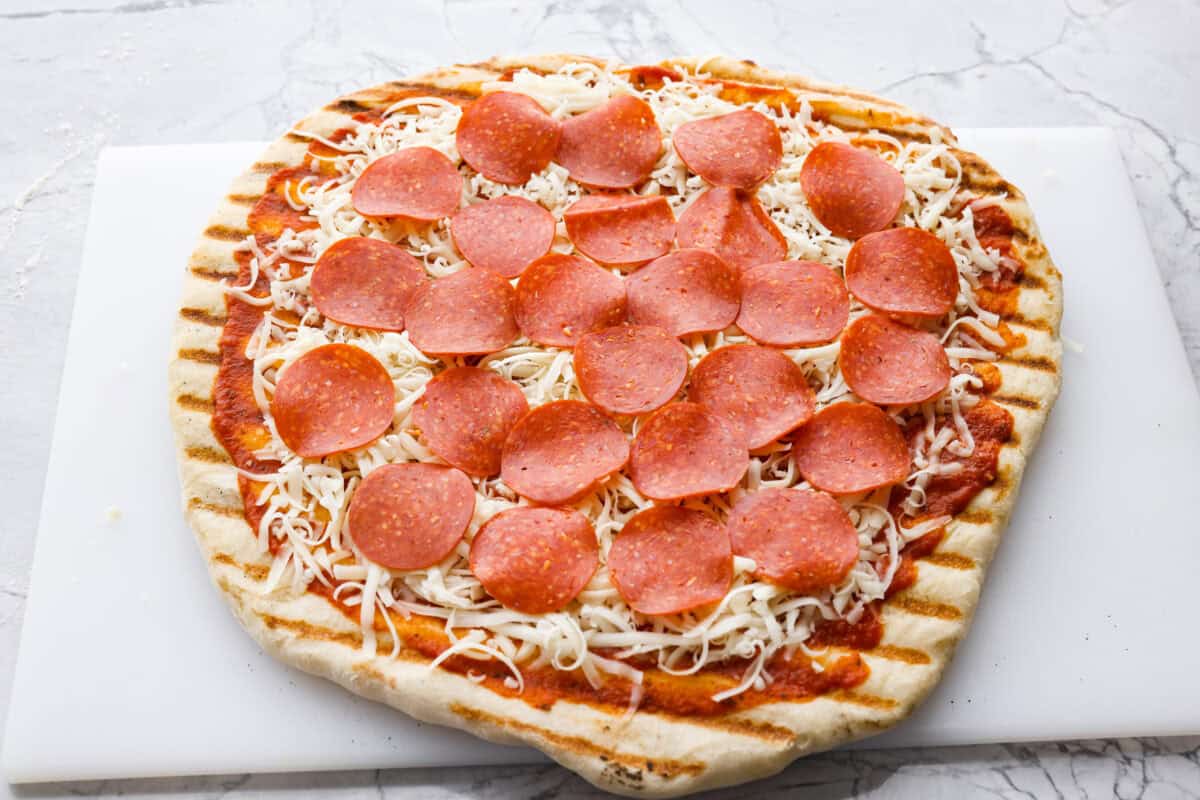 Partially grilled pizza dough with sauce, cheese and pepperoni on it. 