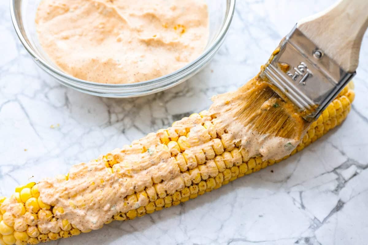 Someone smothering the elote sauce on the cooked corn. 