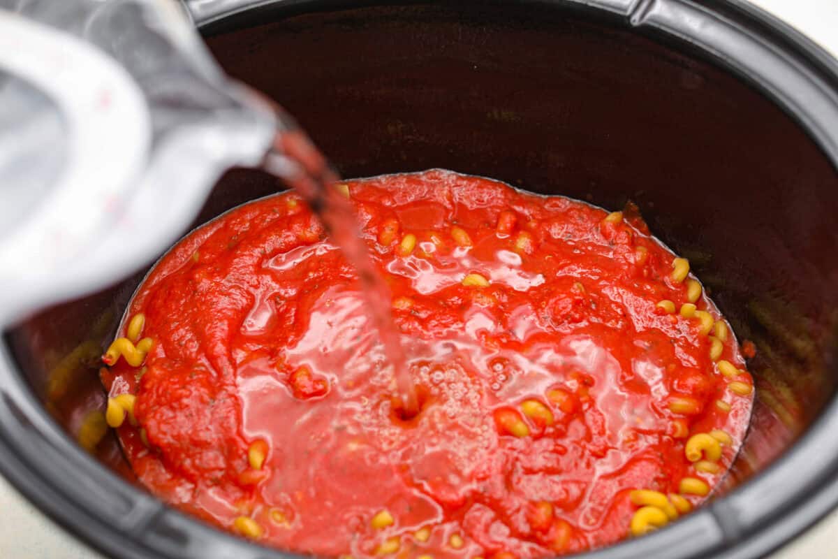 Someone pouring water over the pasta sauce and pasta in the crockpot.