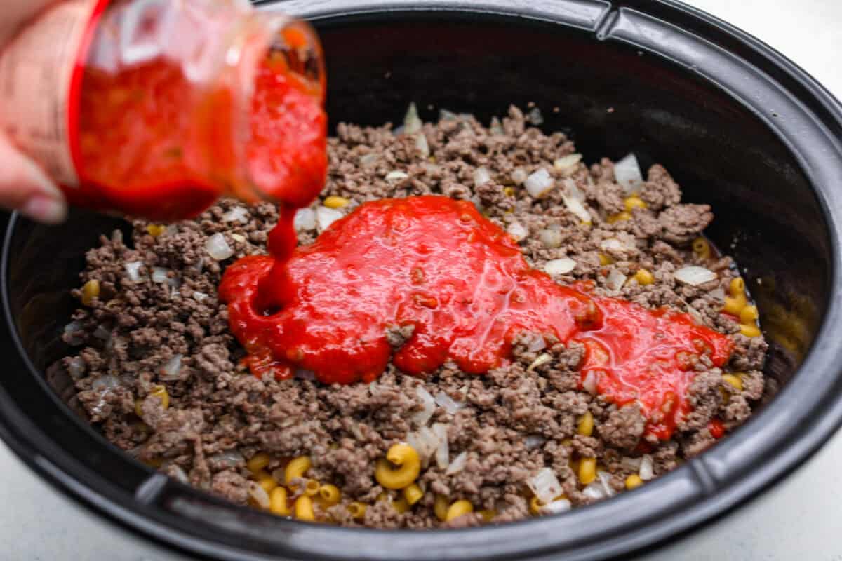Someone pouring a jar of pasta sauce on top of the ground beef and onion mixture. 