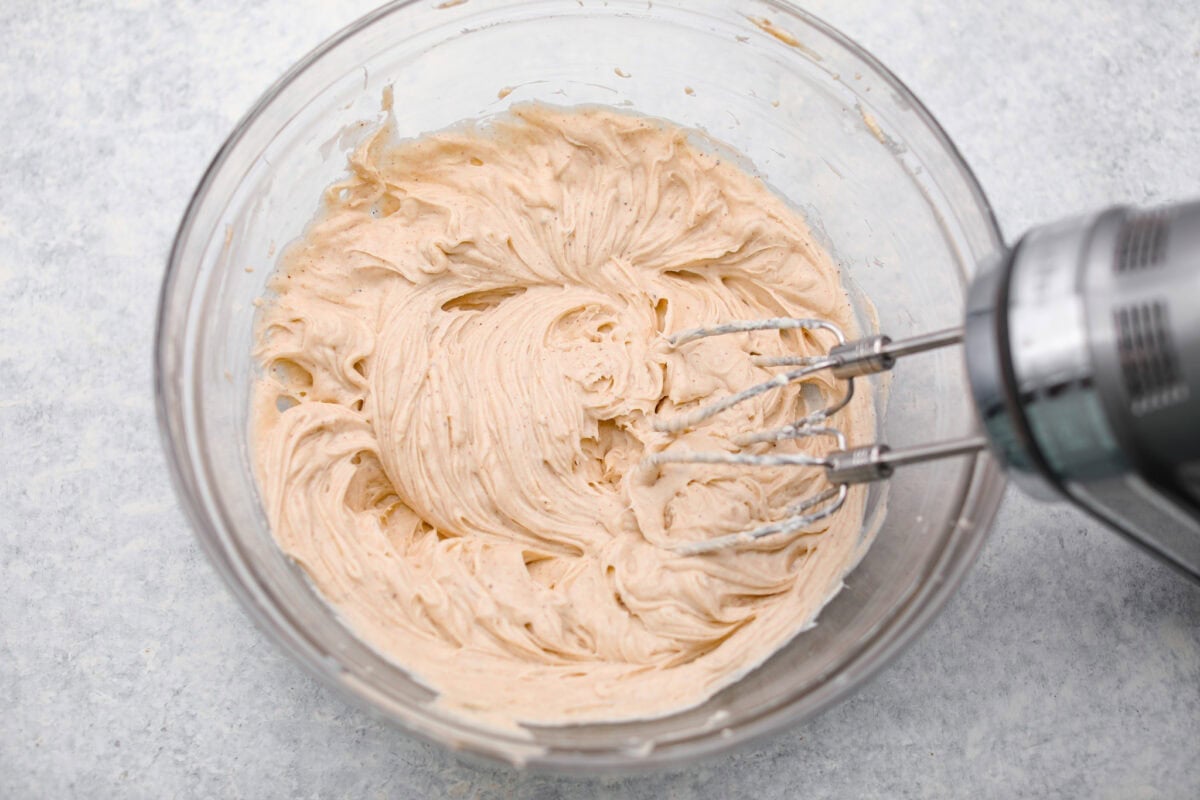 Cream cheese, sugar, vanilla, and seasonings added to a bowl and are being mixed together with a hand mixer.