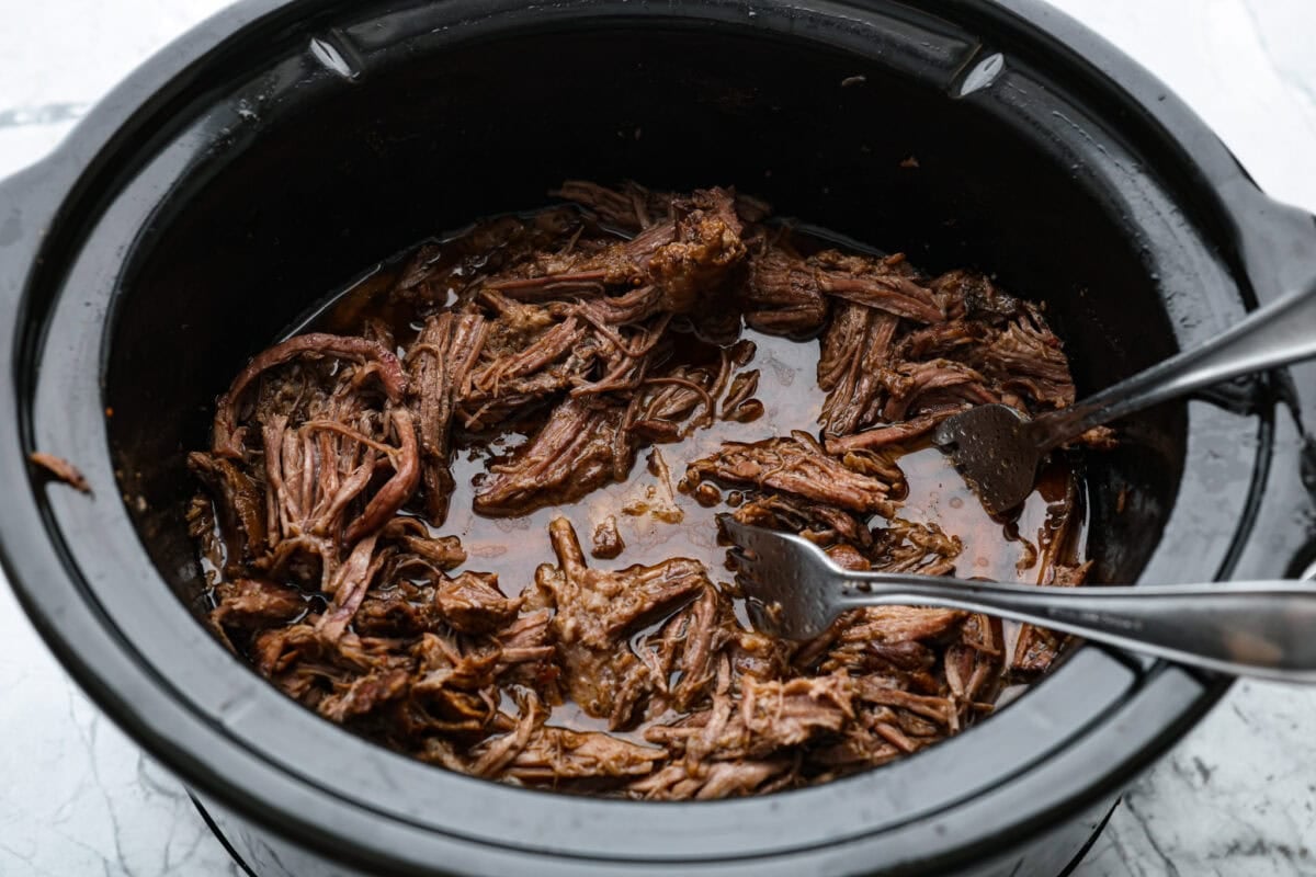 Someone breaking apart the beef with forks in the slow cooker. 