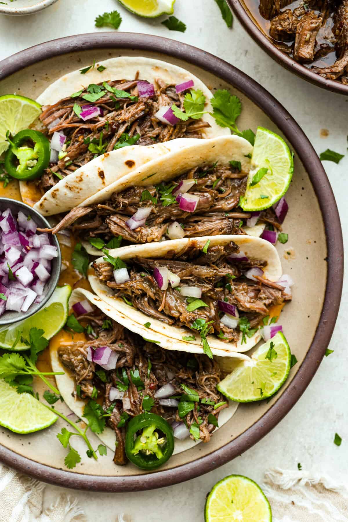 Platter of tacos with slow cooker Barbacoa beef in them and garnished with red onion, cilantro, lime, and jalapeños. 