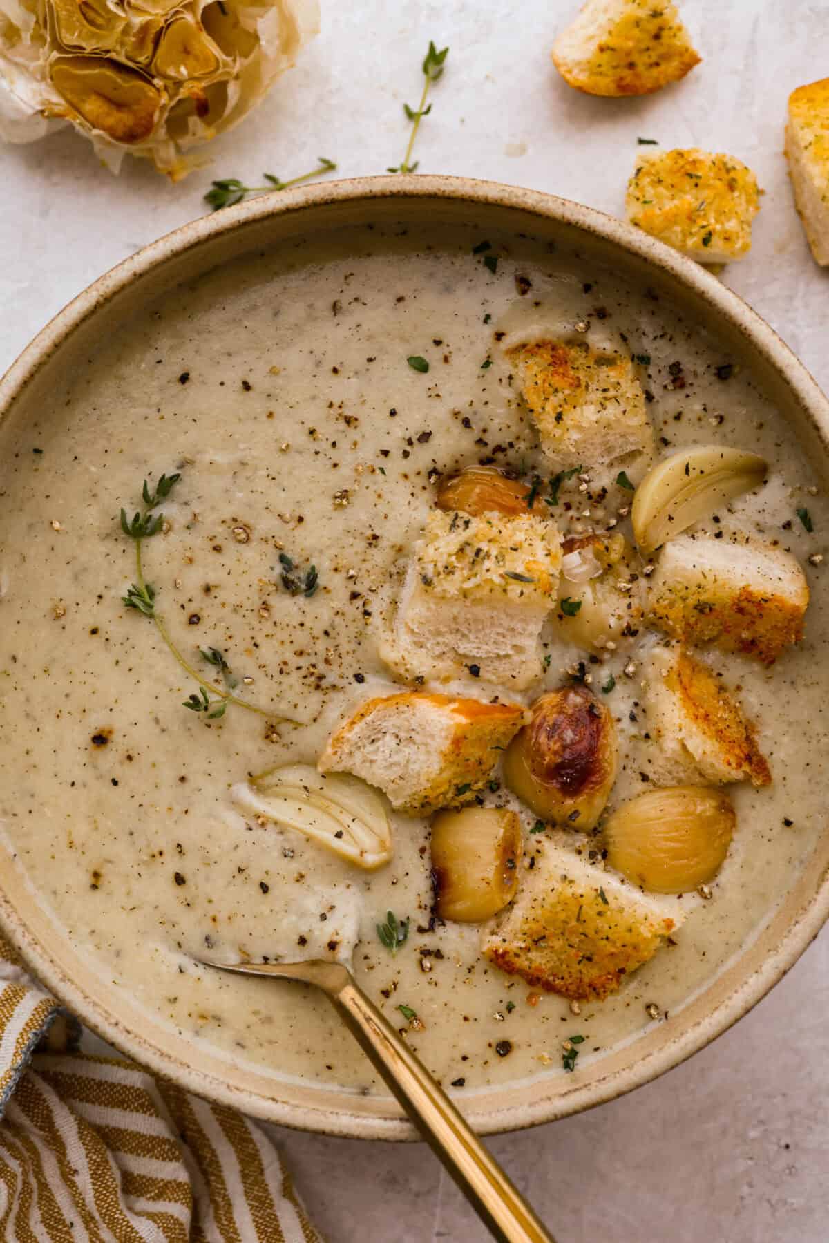 Creamy roasted garlic soup is comfort food with a side of obsession. Combining roasted garlic, tender potatoes, and nutty Parmesan into a cozy soup is absolute perfection. Just make sure you serve it to anyone within hugging distance. Bowl of roasted garlic soup toped with croutons, garlic cloves and pepper.