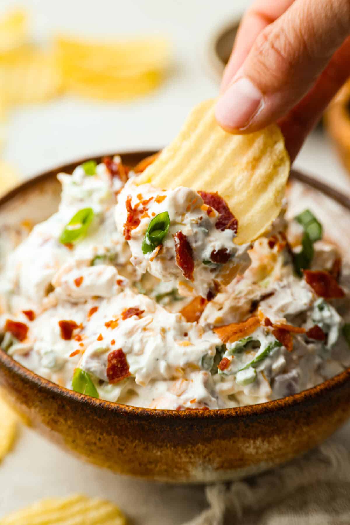 Someone using a ruffled chip to take a scoop of loaded baked potato dip.