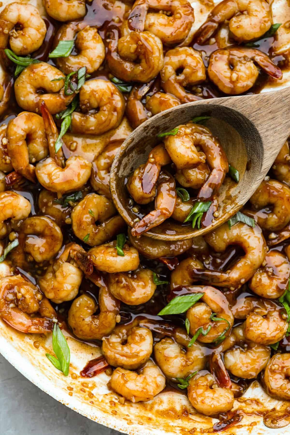 Someone using a wooden spoon to take a scoop of bourbon shrimp out of the skillet. 
