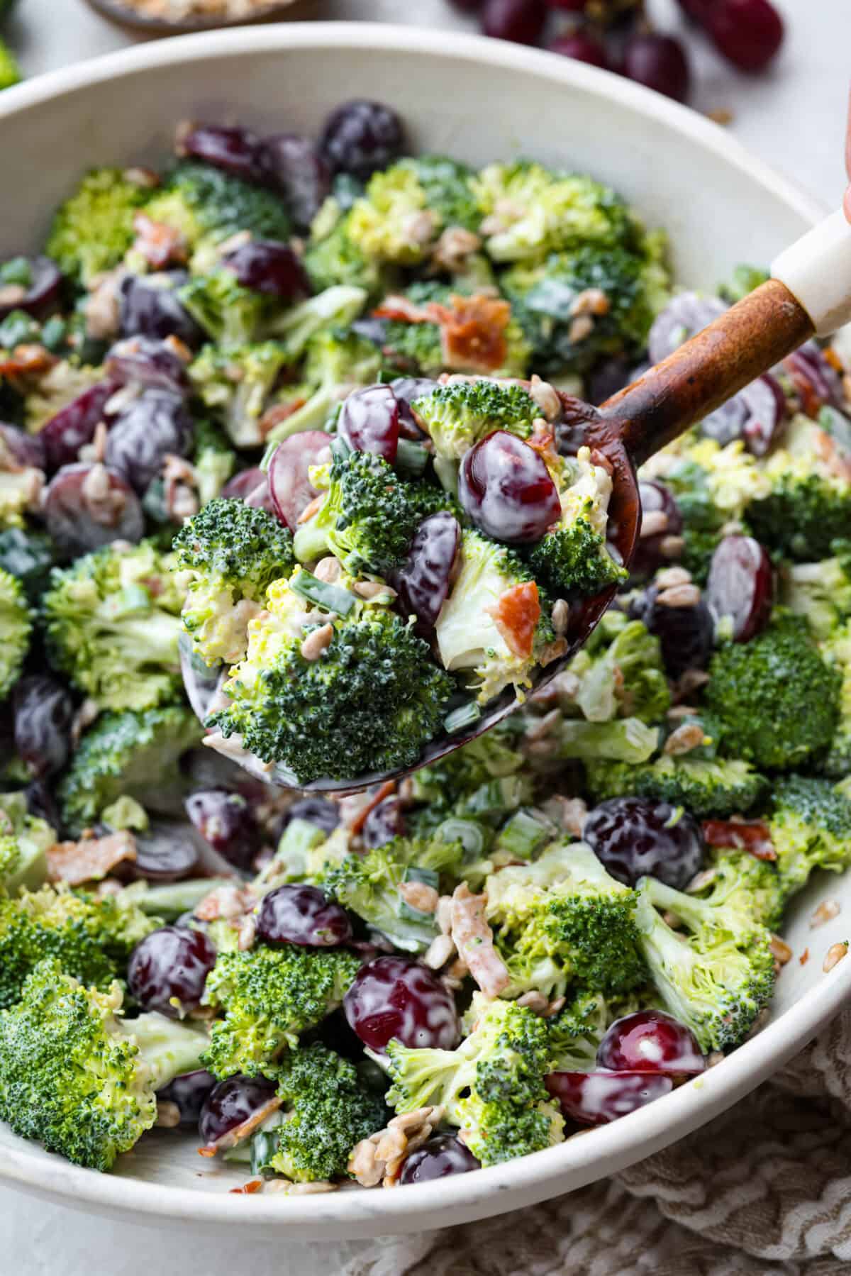 Broccoli grape salad is the crunchy, juicy, smoky combo nobody expects but everyone loves. Sweet grapes, crisp broccoli, smoky bacon, and a tangy lemon dressing come together in just 20 minutes! Someone lifting out a scoop of broccoli grape salad from the large serving bowl.