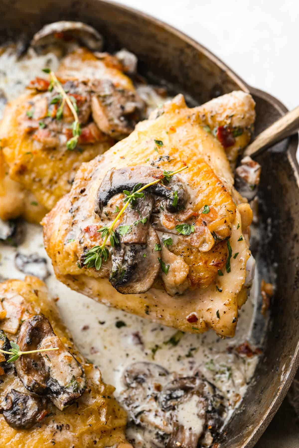 If cozy dinners are your thing, this creamy bacon mushroom thyme chicken needs to happen soon. Between the crispy chicken skin, smoky bacon, and creamy mushroom sauce, every bite tastes rich and savory. Perfect for spooning over mashed potatoes! Someone lifting out a bacon mushroom thyme chicken thigh out of the skillet.