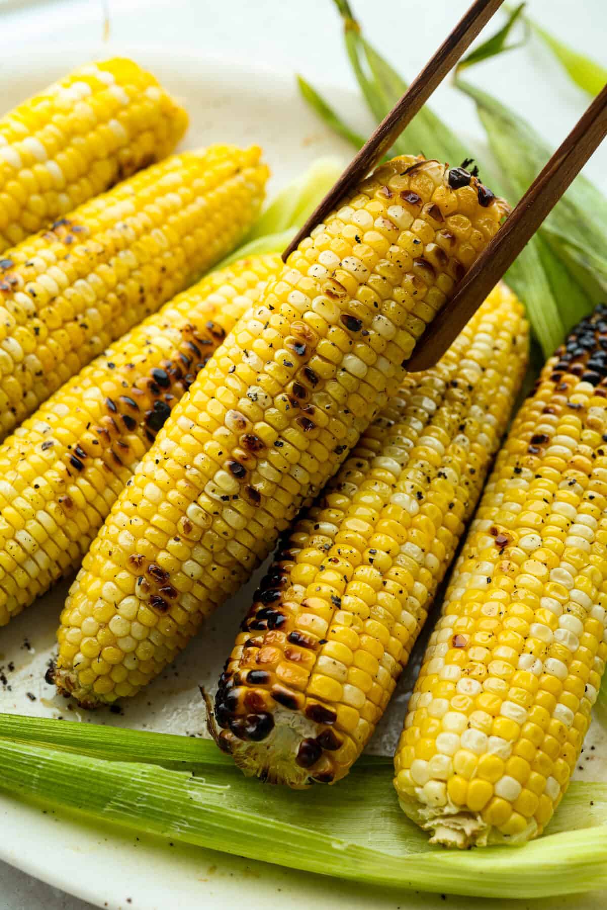 Someone lifting a grilled corn cob off a plate of grilled corn. 