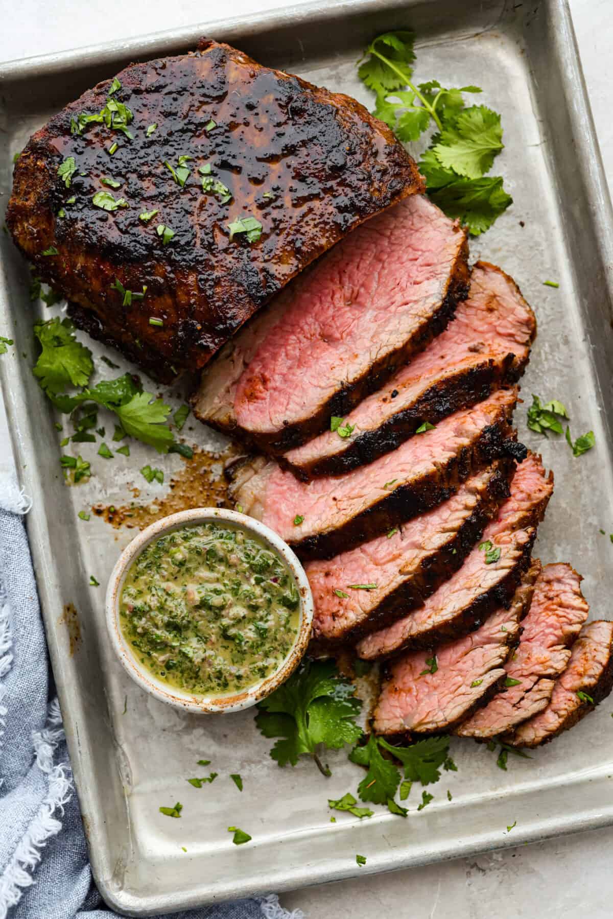 Santa Maria grilled tri-tip is juicy, smoky, and loaded with bold flavor from a delicious spice rub. Grilled until perfectly tender with a gorgeous crust, it’s an easy recipe that’s always a hit at cookouts and summer dinners. Sant Maria grilled tri-tip sliced on a baking sheet next to a small bowl of fresh chimichurri sauce.