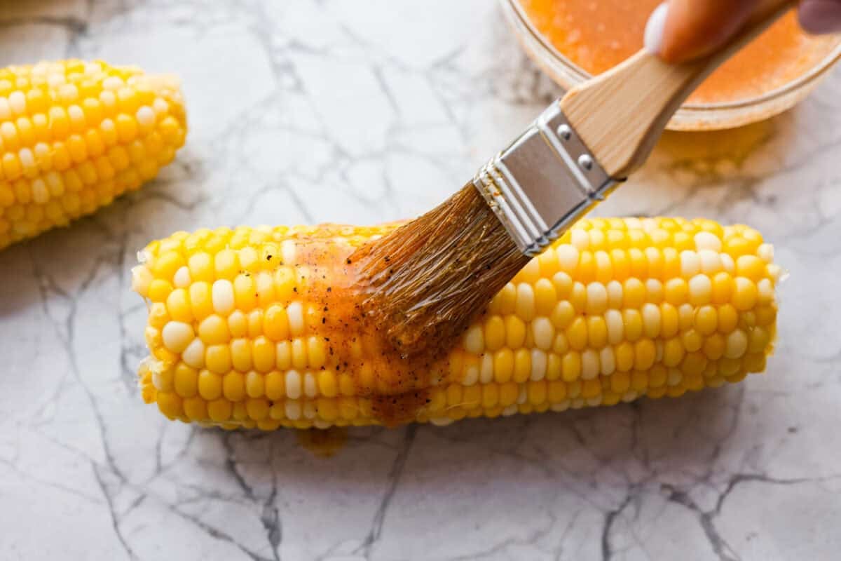 You already know I love a good corn recipe, and this Cajun corn on the cob is my new summer BBQ obsession. It takes sweet, juicy corn and gives it the buttery, spicy glow-up it deserves. Someone basting a cob of corn with the seasoned butter.