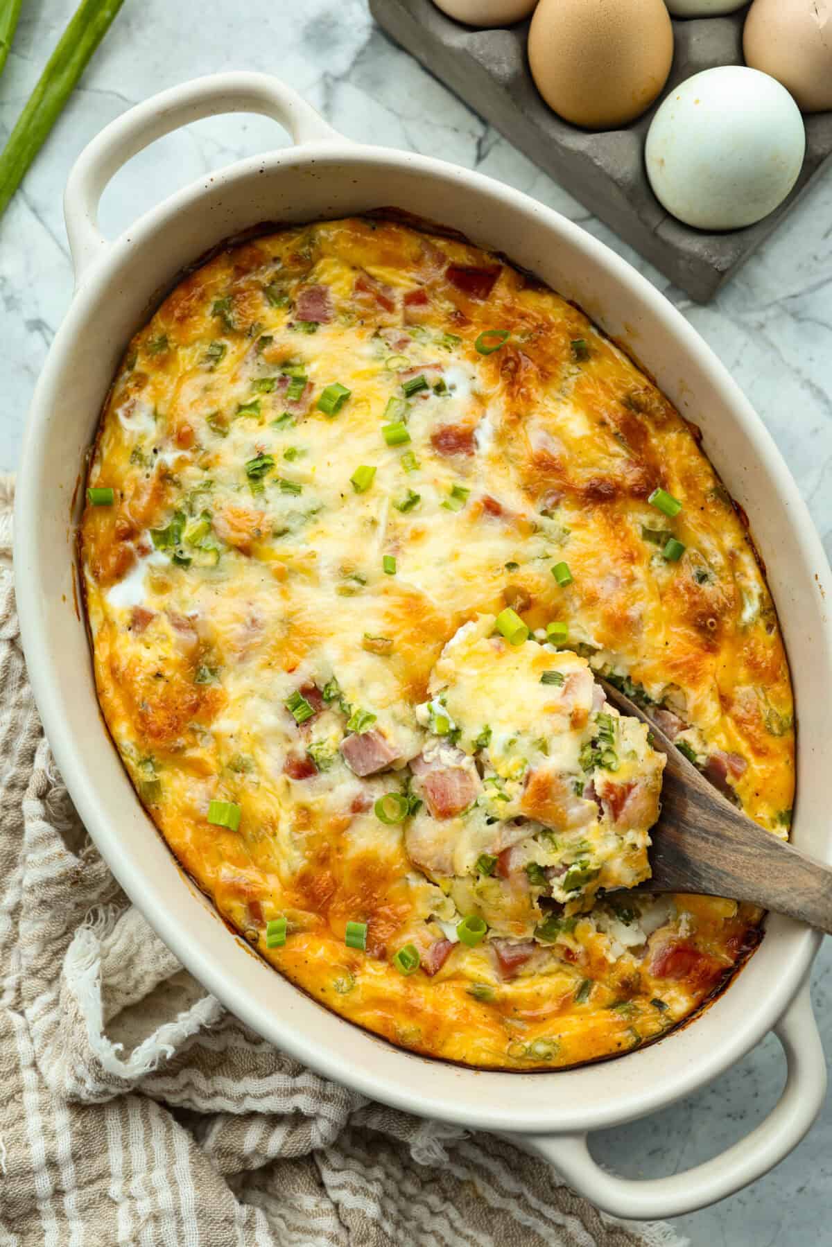 This farmer's casserole is my answer to summer mornings when the kids want cereal again, but I want them to eat something that will actually keep them full. It’s packed with protein, and it's easy to customize with fresh garden veggies. Baked farmer's casserole with a wooden spoon scooping out a serving.