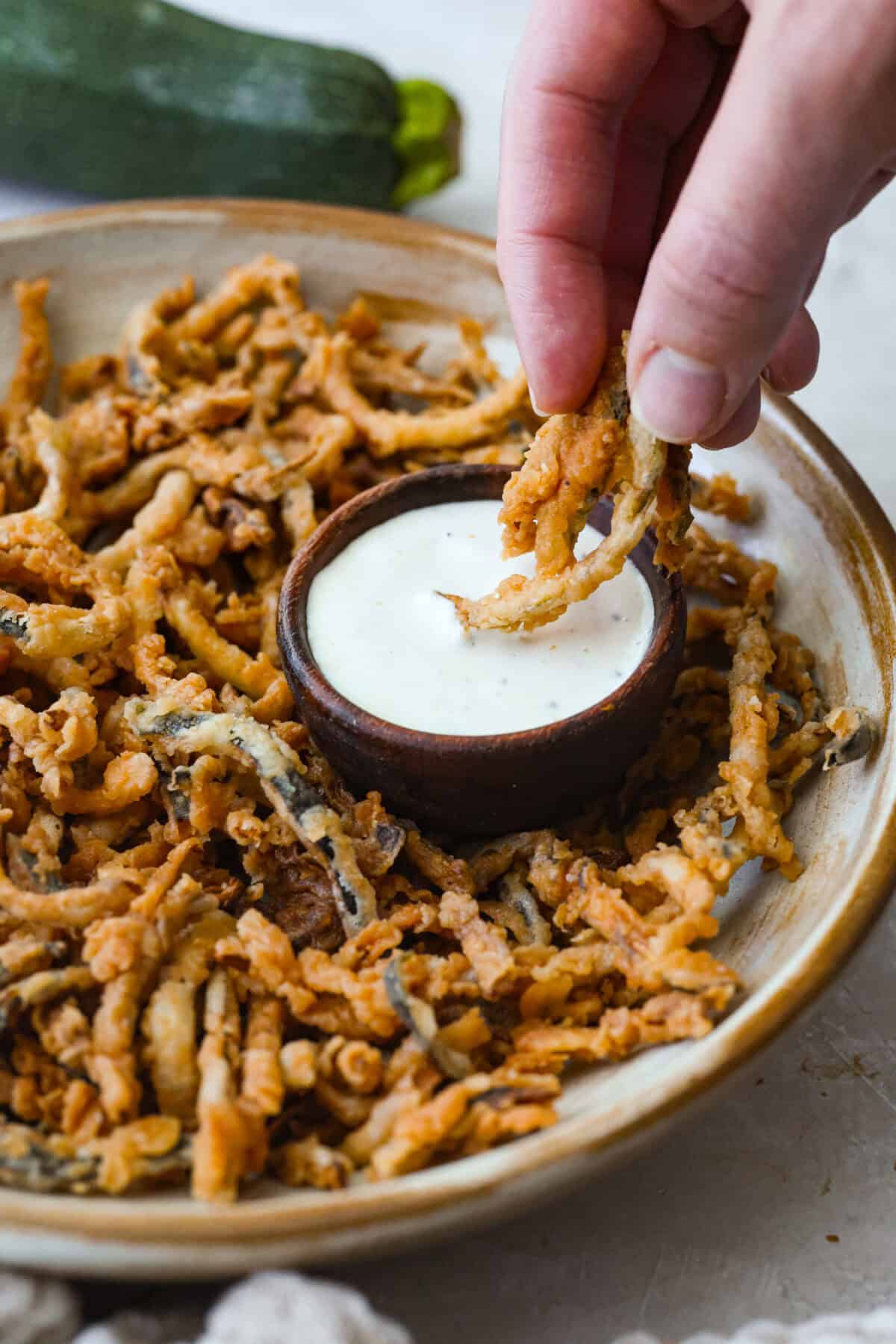 These crispy zucchini strings are golden, crunchy, and seriously hard to stop eating. Lightly battered and fried until perfectly crisp, they turn simple zucchini into an addictive snack or side dish that everyone will reach for first. Someone dipping zucchini strings in ranch dressing.