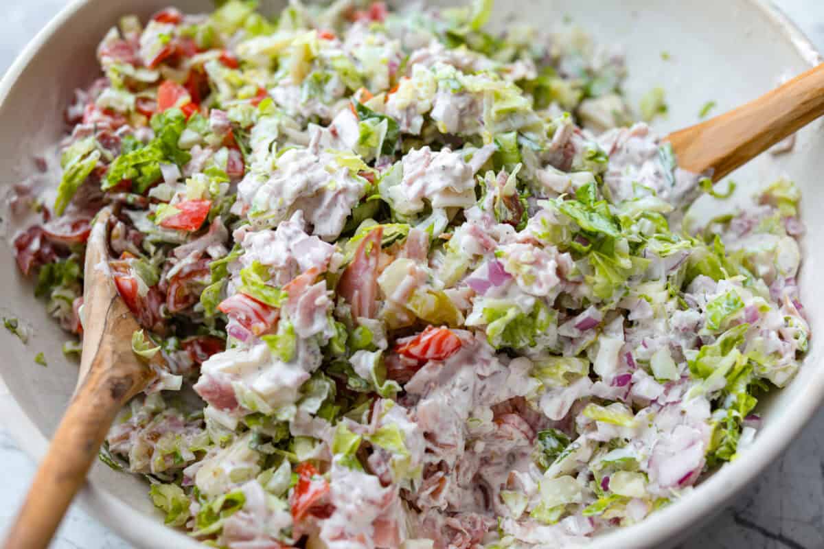 If you're a fan of deli sandwiches, you will love this sub in a tub dip! Made with deli meats, cheese, crisp veggies, and a creamy tangy dressing, it tastes just like your favorite sandwich shop sub in dip form! Someone mixing with the ingredients.