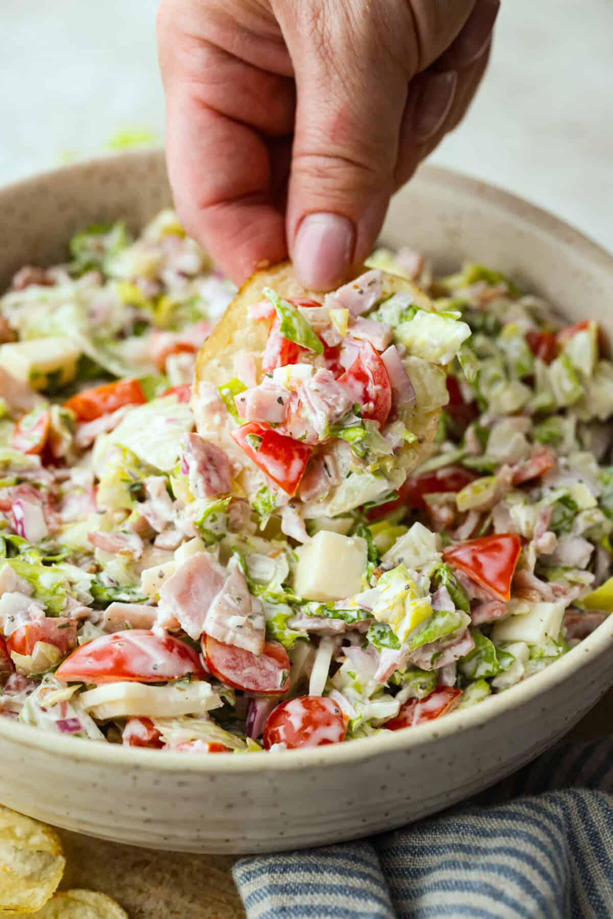 If you're a fan of deli sandwiches, you will love this sub in a tub dip! Made with deli meats, cheese, crisp veggies, and a creamy tangy dressing, it tastes just like your favorite sandwich shop sub in dip form! Someone scooping a bite of the sub in a tub dip with a potato chip.