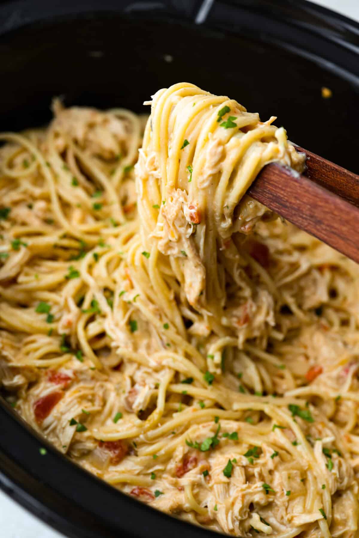 This crockpot chicken spaghetti will have everyone running to the dinner table! The slow cooker does the work while the chicken becomes tender and flavorful, then it's tossed with spaghetti, melty cheese, and a creamy sauce. Someone lifting up a scoop of crockpot chicken spaghetti with wooden tongs.