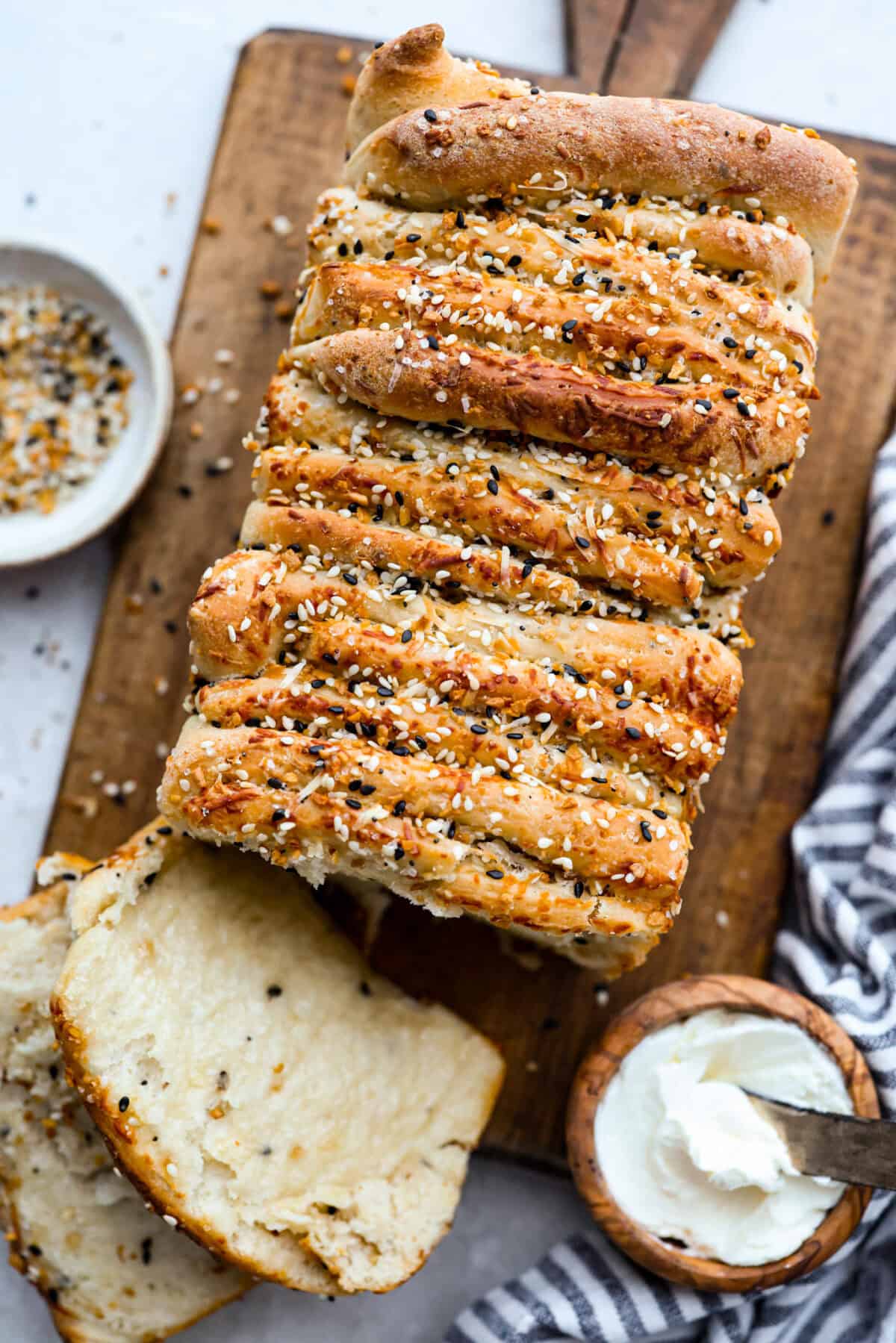 Everything bagel pull apart bread is soft, cheesy, and loaded with savory flavor in every layer. The homemade dough is stacked with Parmesan cheese and everything bagel seasoning, then baked until golden and fluffy. Loaf of everything bagel pull apart bread on a cutting board, sliced with a small bowl of cream cheese next to it.