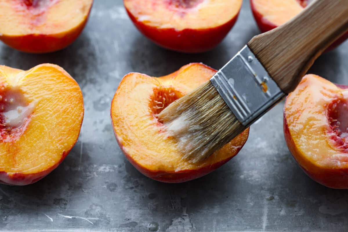 Grilled peaches are one of my favorite ways to turn ripe summer fruit into an easy dessert. The grill brings out their natural sweetness and adds just a little smoky flavor. Serve them warm with vanilla ice cream for a simple treat that feels extra special. Someone using a basting brush to brush on the melted butter.