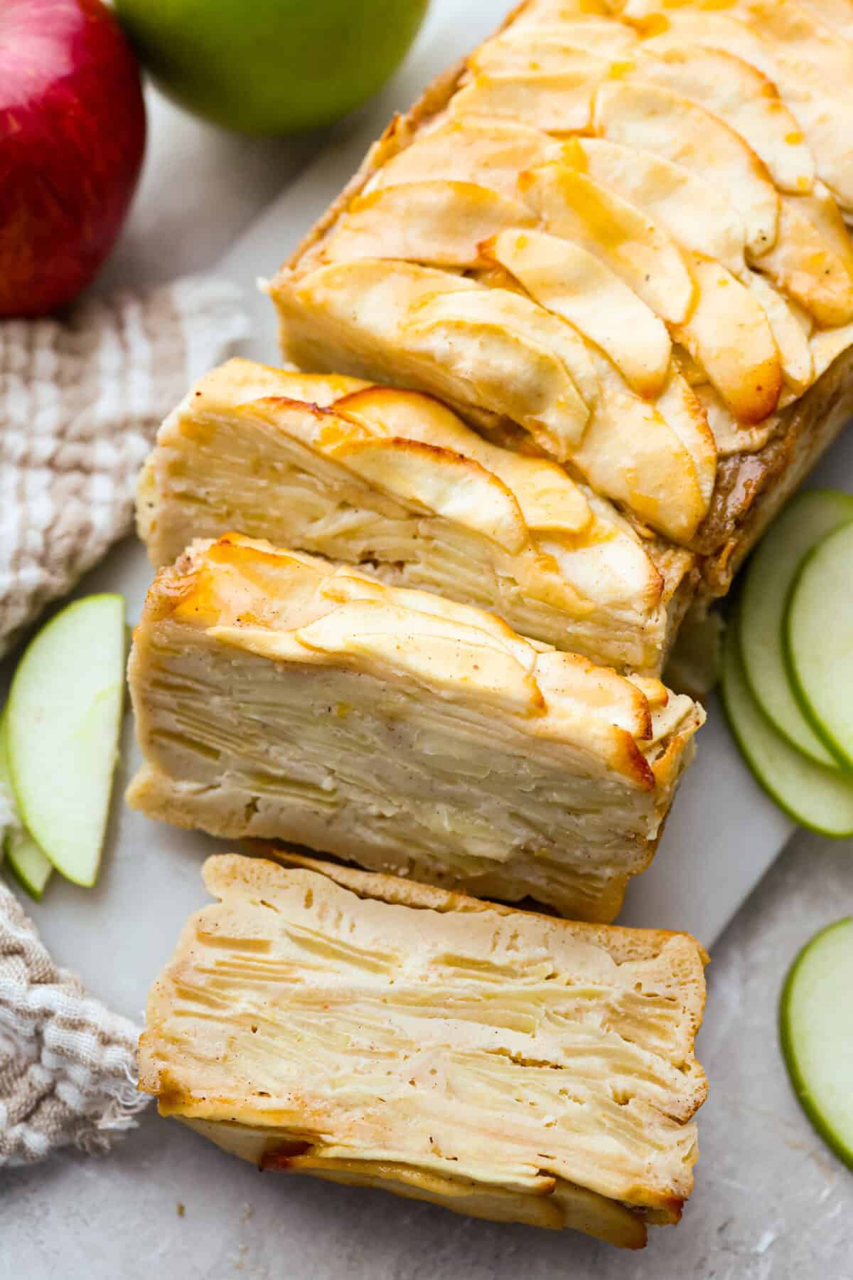 This invisible apple cake is a little magical! Paper-thin apple slices are coated in a simple, lightly spiced batter, then layered in a loaf pan and baked into a tender, custardy cake. Such a delicious way to enjoy fresh apples! Invisible Apple Cake with a few slices cut out of it on a cutting board surrounded by apples.