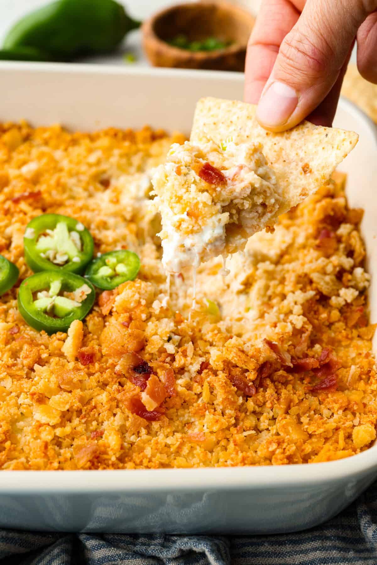 If you love jalapeño poppers, you need this dip. It’s creamy, cheesy, loaded with bacon, and topped with buttery Ritz crackers. Best of all, it takes just 10 minutes to prep, and is perfect for game day! Someone using a chip to scoop some of the jalapeño popper dip out of the baking dish.