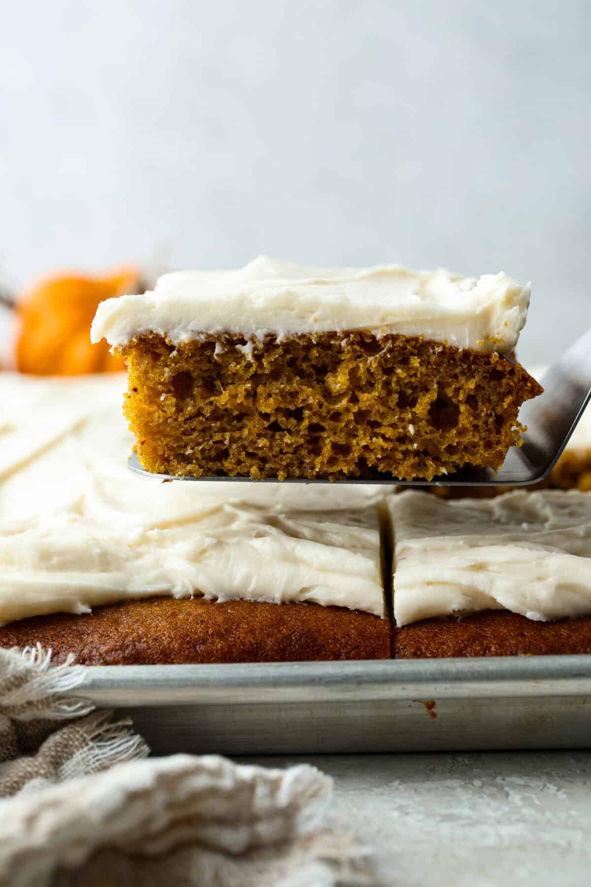 Pumpkin Sheet Cake Bars are one of my oldest son’s favorite desserts, and honestly, I get it! They’re incredibly moist and fluffy, full of warm pumpkin spice, and topped with the best cream cheese frosting. Someone lifting a slice of pumpkin sheet cake bar out of the pan.