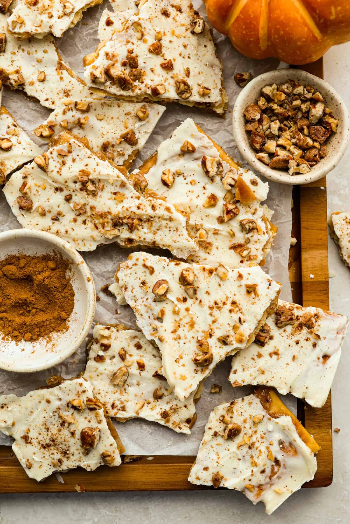 Pumpkin spice crack is Christmas crack with a fall twist! Buttery toffee, salty crackers, white chocolate, pumpkin pie spice, and crunchy praline pecans make this an easy treat you won’t be able to stop snacking on. Top down shot of pumpkin pie spice crack broken up on a cutting board.