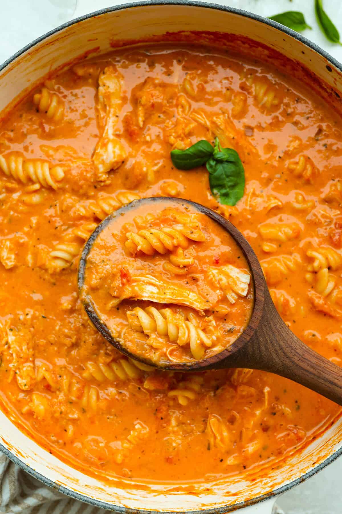 Craving chicken parm? Grab a spoon! This chicken parmesan soup is loaded with tender chicken, pasta, and a rich, creamy tomato broth. Finish with crispy, buttery breadcrumbs for a delicious twist on the Italian classic! Pot of chicken parmesan soup with a wooden ladle and fresh basil leaf.