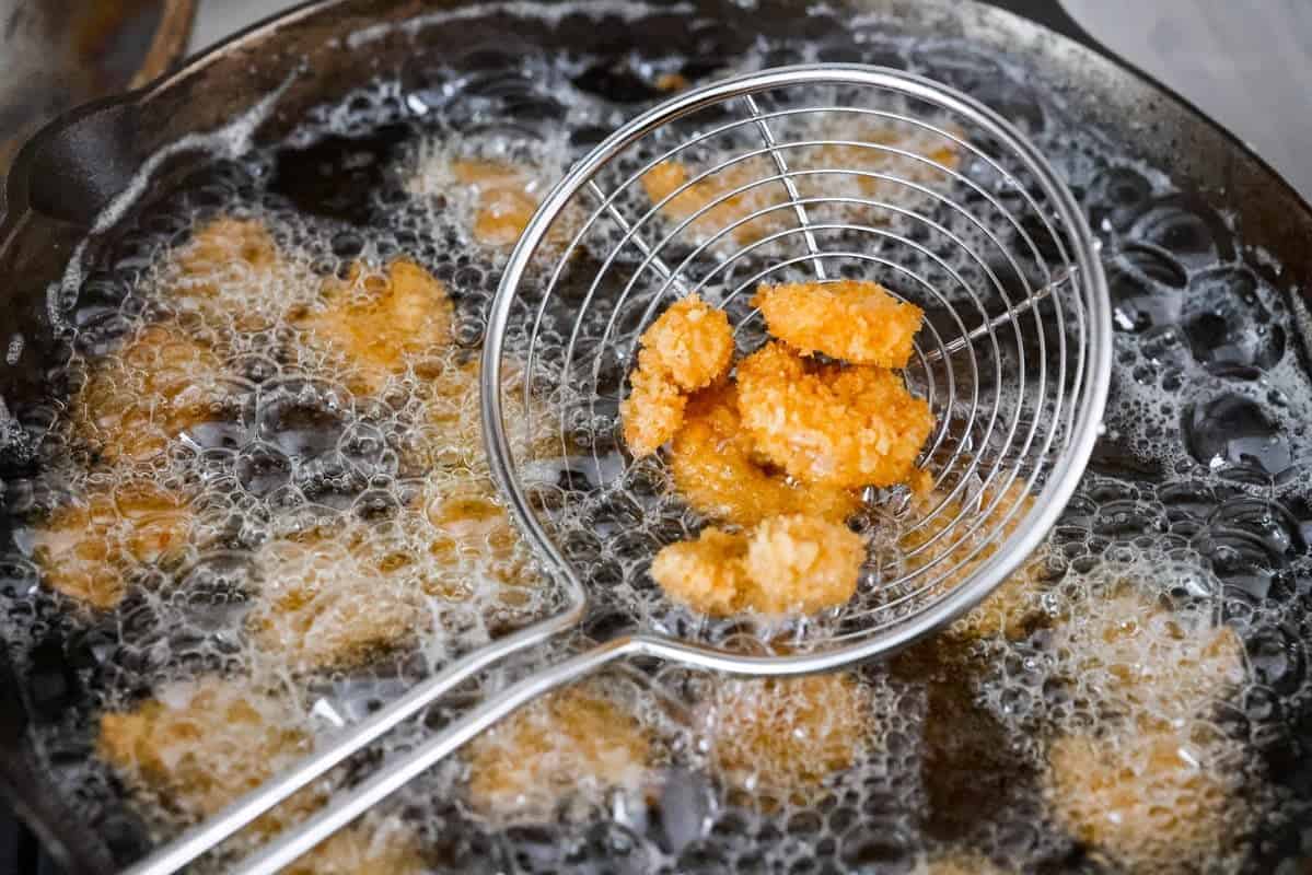 These crispy popcorn shrimp are little bites of golden, crunchy goodness! Juicy shrimp get coated in seasoned panko and fried until perfectly crisp. Serve them hot with a squeeze of lemon and cocktail sauce for dipping! Someone lifting out fried shrimp out of the oil.