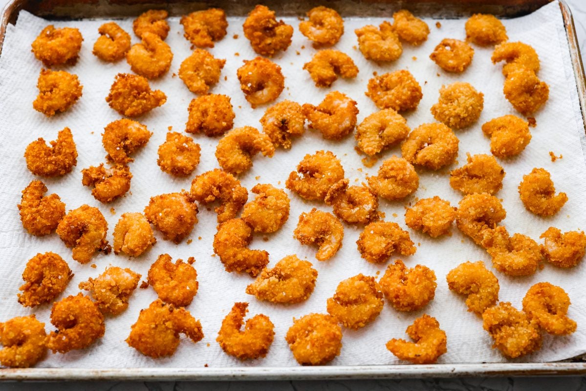 These crispy popcorn shrimp are little bites of golden, crunchy goodness! Juicy shrimp get coated in seasoned panko and fried until perfectly crisp. Serve them hot with a squeeze of lemon and cocktail sauce for dipping! Crispy popcorn shrimp spread out on a paper towel after being fried.
