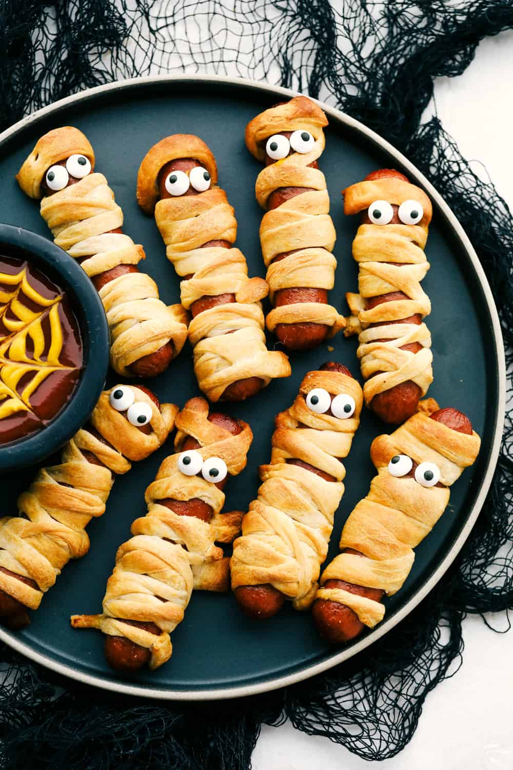 Nothing screams fun, quite like Mummy Hot Dogs for Halloween dinner. Serve these fun little vittles before trick or treating or at your next Halloween party. Mummy Hot dogs on a plate with spider web dip.