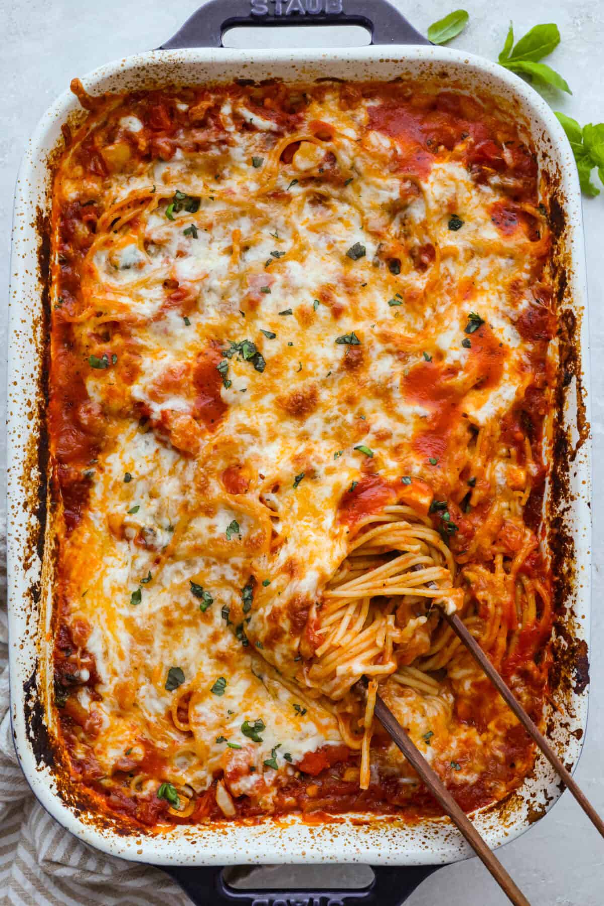 This no boil spaghetti bake makes pasta night SO easy! Everything goes straight into one baking dish, uncooked spaghetti included, then bakes under a layer of melty mozzarella for a cozy, cheesy dinner with hardly any prep. Top down shot of the baked no boil pasta bake with some wooden tongs getting a scoop.
