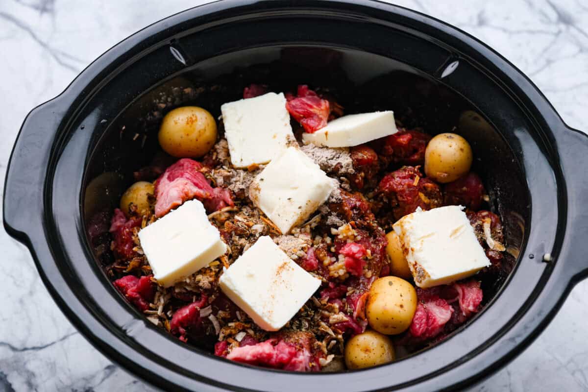 Slow cooker with meat, potatoes, seasoning packets, broth, and butter.